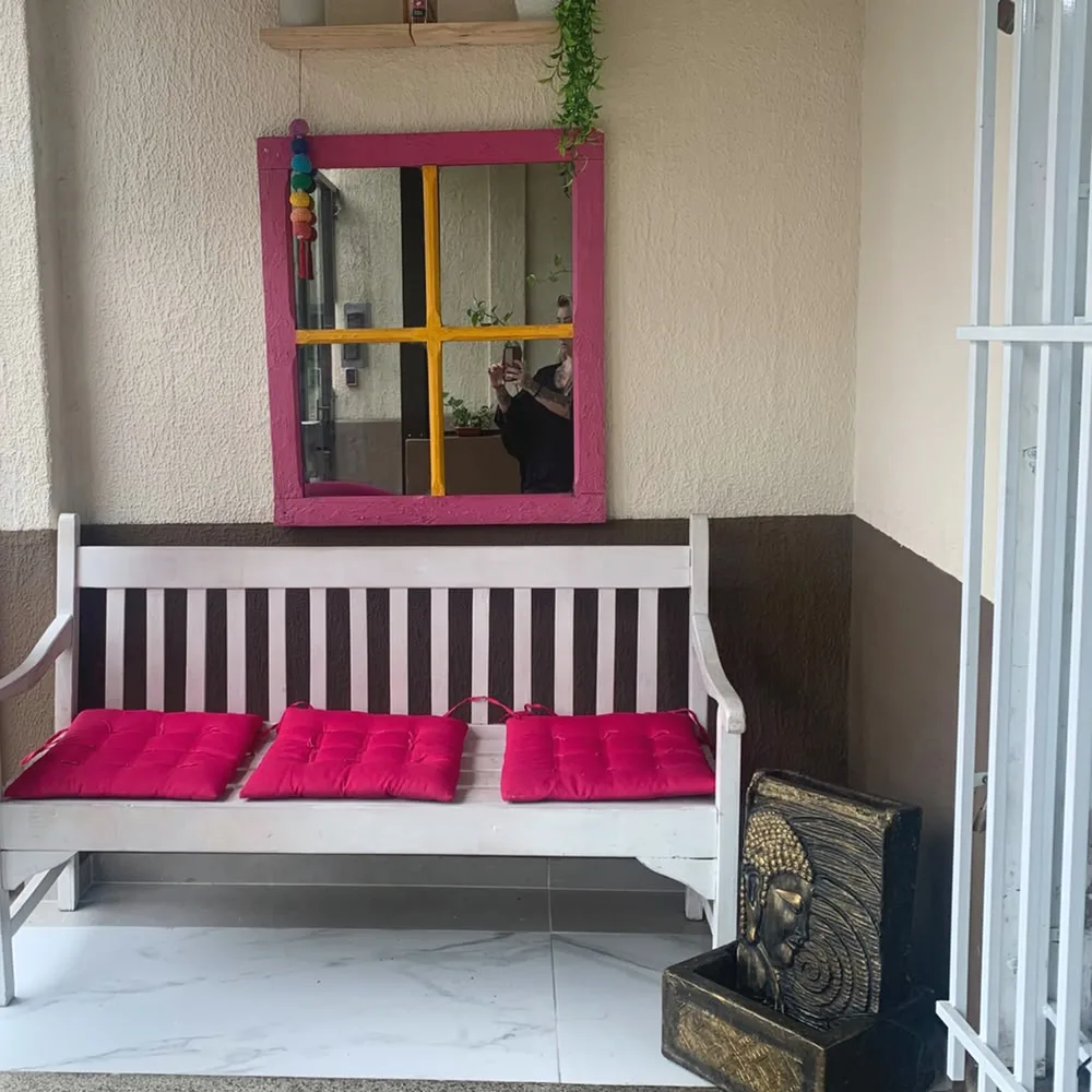 O banco branco com almofadas pink e o espelho de moldura colorida, na entrada